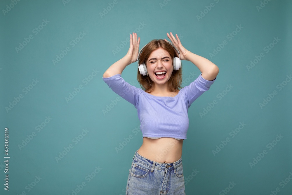 Charming happy smiling young blonde woman wearing blue crop top isolated on blue background with copy space for text wearing white wireless bluetooth headphones listening to good music dancing and Charming happy smiling young blonde woman wearing blue crop top isolated on blue background with copy space for text wearing white wireless bluetooth headphones listening to good music dancing and