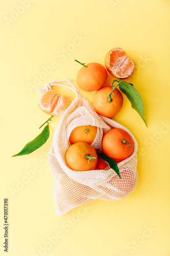 Fresh clementines with leaves in reusable string bag on yellow background, top view
