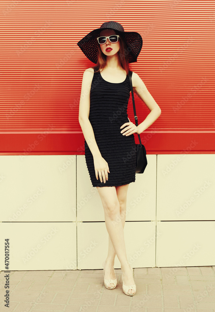 Portrait of beautiful young woman with handbag wearing a black summer hat and dress on red background Portrait of beautiful young woman with handbag wearing a black summer hat and dress on red background