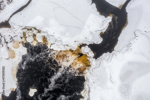 Top view of the river waterfall. Winter aerial view of a freezing river. Animal tracks on the ice covered with snow. Yellow peat water. Cold winter weather. Tosna rapids, Leningrad region, Russia. photo