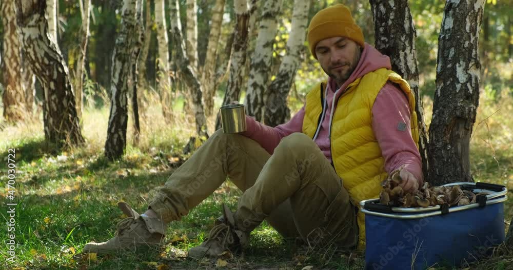 a man in a yellow vest and a hat sits in the autumn forest with a cup of hot tea. young male mushroom picker with a large basket looks for, collects mushrooms in the forest. Mushroom picking in season a man in a yellow vest and a hat sits in the autumn forest with a cup of hot tea. young male mushroom picker with a large basket looks for, collects mushrooms in the forest. Mushroom picking in season
