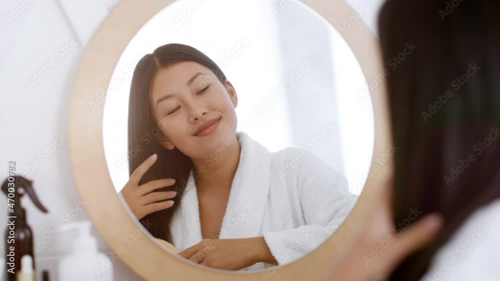 Hair Care concept. Young cheerful asian woman brushing her hair, looking at mirror at bathroom and smiling Hair Care concept. Young cheerful asian woman brushing her hair, looking at mirror at bathroom and smiling