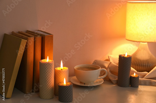 Burning candles, cup of coffee, books and glowing lamp on table near wall
