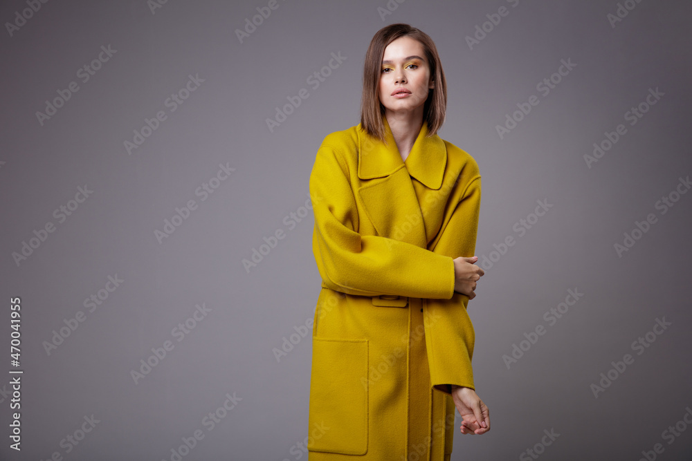 High fashion photo of a beautiful elegant young woman in a pretty yellow mustard coat, black ankle boots posing over gray background. Slim figure. Bob haircut. Studio Shot.