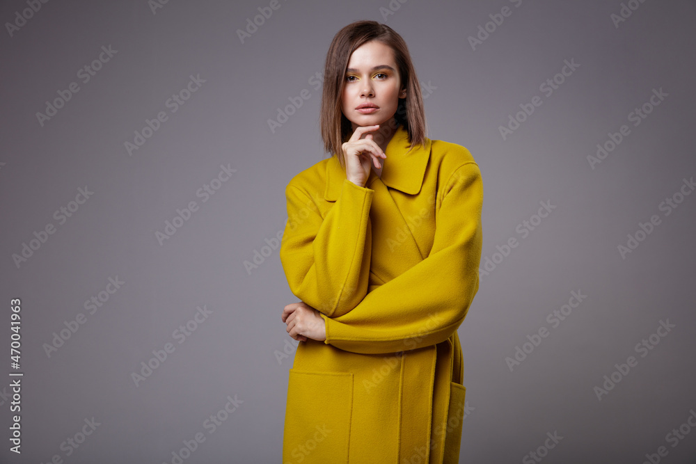 High fashion photo of a beautiful elegant young woman in a pretty yellow mustard coat, black ankle boots posing over gray background. Slim figure. Bob haircut. Studio Shot. High fashion photo of a beautiful elegant young woman in a pretty yellow mustard coat, black ankle boots posing over gray background. Slim figure. Bob haircut. Studio Shot.
