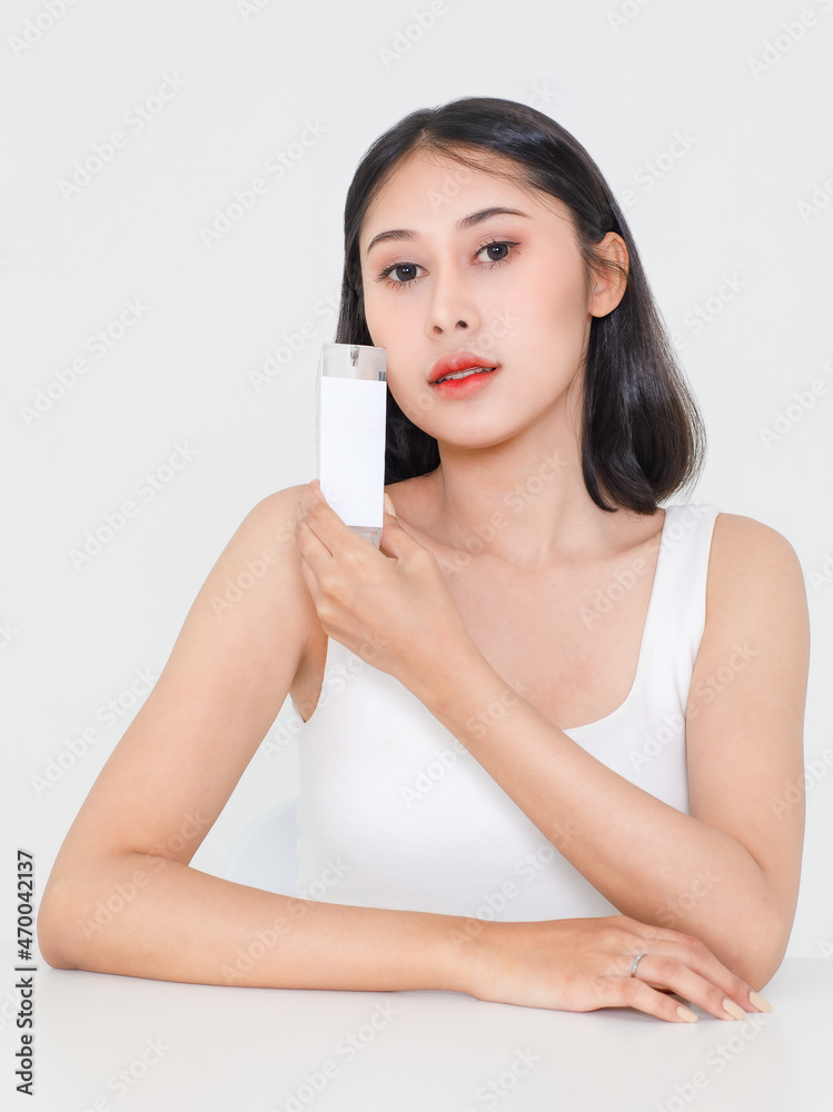 Portrait beauty studio shot millennial Asian black hair sexy female model in tank top singlet look at camera holding showing serum facial moisturizer skincare package near face on white background Portrait beauty studio shot millennial Asian black hair sexy female model in tank top singlet look at camera holding showing serum facial moisturizer skincare package near face on white background