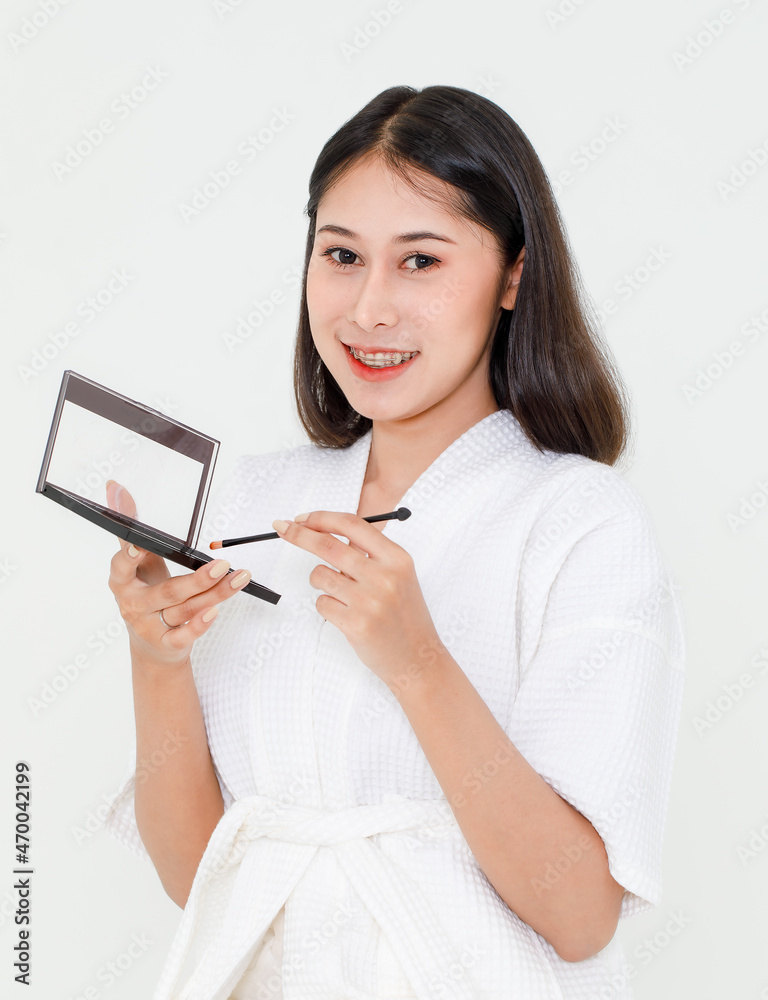 Portrait beauty shot of millennial young Asian female model wearing makeup in shower rope standing smiling look at camera holding colorful eye shadow with mirror and brush on white background Portrait beauty shot of millennial young Asian female model wearing makeup in shower rope standing smiling look at camera holding colorful eye shadow with mirror and brush on white background