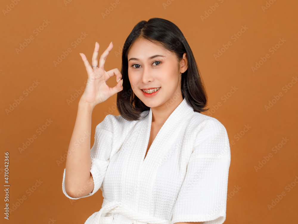 Portrait closeup studio shot of millennial Asian black short hair female model wearing makeup in white shower rope standing smiling look at camera posing holding ok hand sign up on brown background Portrait closeup studio shot of millennial Asian black short hair female model wearing makeup in white shower rope standing smiling look at camera posing holding ok hand sign up on brown background