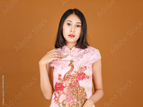 Portrait closeup studio shot millennial Asian female model in pink Chinese cheongsam qipao traditional peacock & flowers pattern dress look at camera holding hand slick hair back on brown background