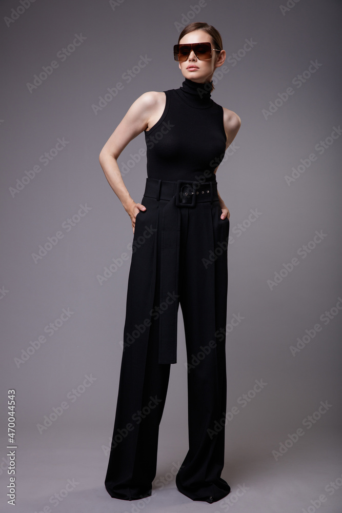 High fashion photo of a beautiful elegant young woman in a pretty black pants, sleeveless top, stylish sunglasses posing over gray background. Slim figure. Studio Shot. High fashion photo of a beautiful elegant young woman in a pretty black pants, sleeveless top, stylish sunglasses posing over gray background. Slim figure. Studio Shot.