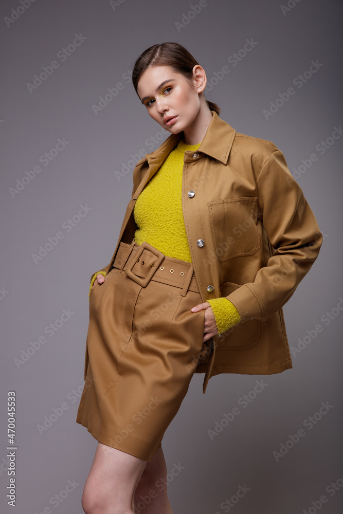 High fashion photo of a beautiful elegant young woman in a pretty brown beige sand color jacket, skirt, yellow mustard sweater posing over gray background. Slim figure. Collected hair. Studio Shot High fashion photo of a beautiful elegant young woman in a pretty brown beige sand color jacket, skirt, yellow mustard sweater posing over gray background. Slim figure. Collected hair. Studio Shot
