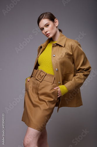 High fashion photo of a beautiful elegant young woman in a pretty brown beige sand color jacket, skirt, yellow mustard sweater posing over gray background. Slim figure. Collected hair. Studio Shot