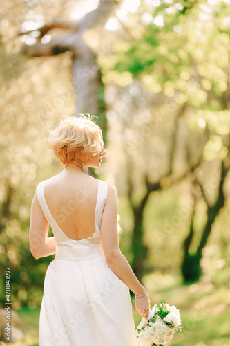 Bride in a white dress with a bouquet of flowers in her hands stands in the park with her head turned to the right