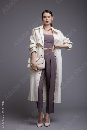 High fashion photo of a beautiful elegant young woman in a pretty white coat, purple top, pants, accessories, handbag posing over gray background. Studio Shot. Slim figure. Make up, hairstyle.