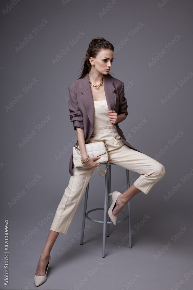 High fashion photo of a beautiful elegant young woman in a pretty purple jacket, white top, pants, accessories, handbag posing on gray background. Studio Shot. Slim figure. Model is sitting on a chair