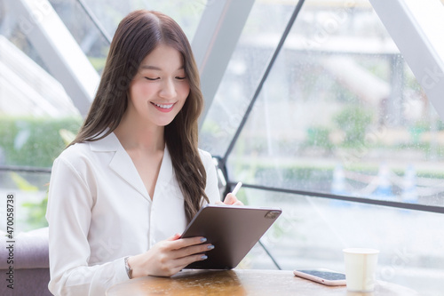 Young Asian business woman is looking at tablet or notepad in her hands while she is sitting at coffee shop among business background in the city.