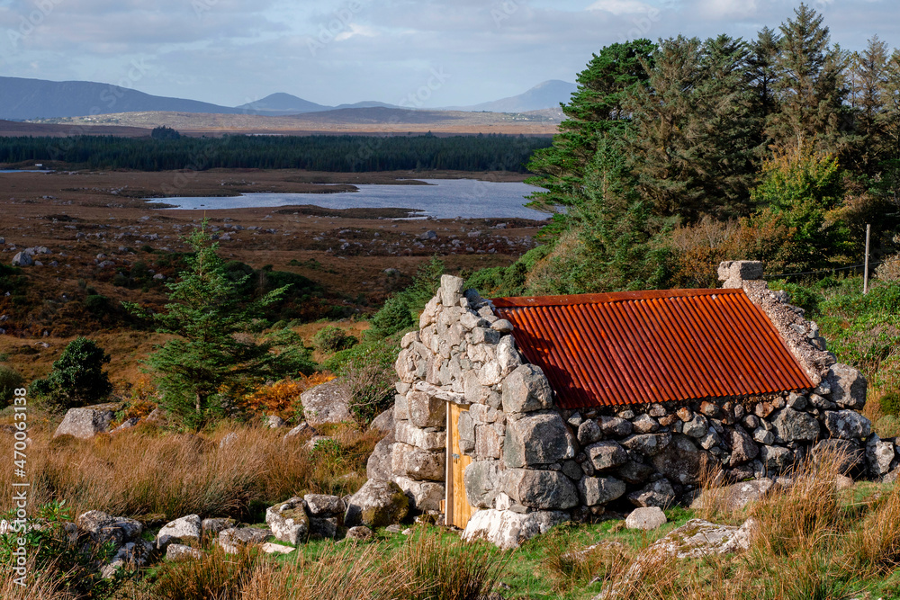 Small stone building with rusty red metal roof with beautiful nature scenery in the background. Connemara area, county Galway, Ireland. Storage hut in a field. Historical building. Cloudy sky