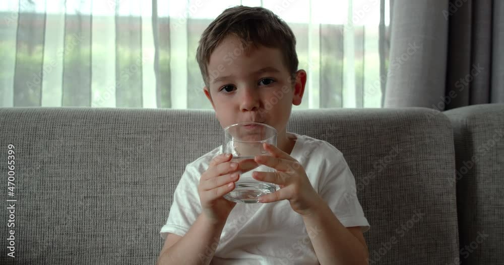 Cute baby boy drinking a glass of water sitting on the couch at home. Slow motion little kid drinking water. Close-up. Portrait Funny little Child is drinking a cup of water. Cute baby boy drinking a glass of water sitting on the couch at home. Slow motion little kid drinking water. Close-up. Portrait Funny little Child is drinking a cup of water.
