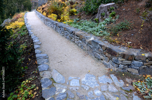 road cut into the slope. above and below the road is a stone dry wall. nature trail through the autumn park with a drain and a metal grid. slopes overgrown with drought perennials yellow, yew, boulder photo