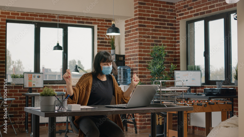 Cheerful woman celebrating successful achievement, feeling happy and dancing at desk, wearing face mask. Accomplished employee reading good news on laptop and enjoying satisfaction