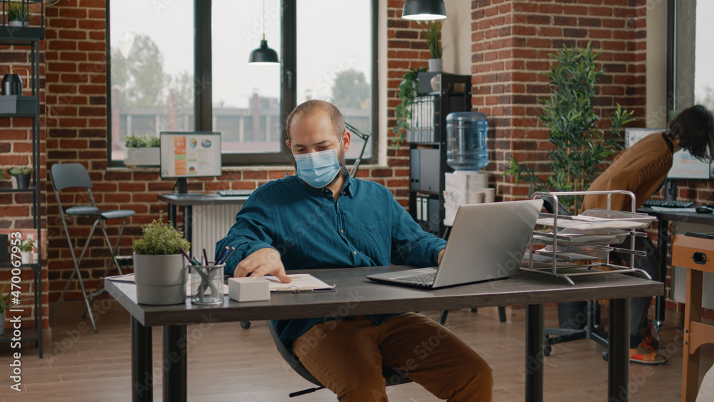 Employee checking data rate charts on papers and laptop, looking at display and wearing face mask. Business man using technology to plan project for presentation and startup meeting