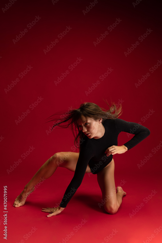 Portrait of emotional young flexible contemp dancer, ballerina dancing isolated on dark red background. Art, beauty, inspiration concept.