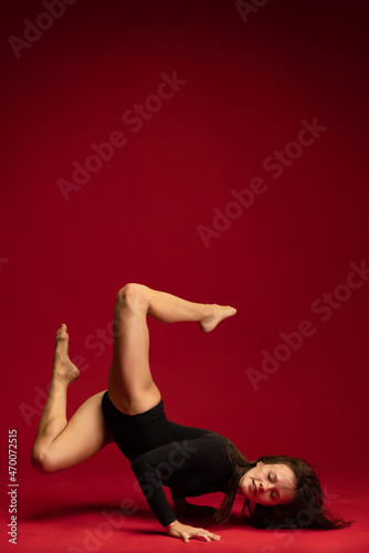 Studio shot of young flexible girl, female contemp dancer, ballerina in action isolated on dark red background. Art, beauty, inspiration concept.