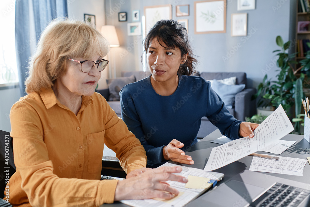 Obraz premium Senior businesswoman in eyeglasses working on laptop with documents at the table together with assistant, they working at home