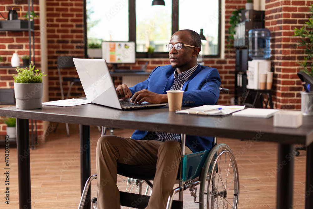 Business man sitting in wheelchair working on laptop to plan project and strategy in office. Person with disability using computer to work on company startup and do management research.