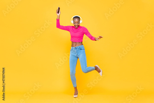 Full length portrait of happy African American woman listening to music and dancing in studio yellow color isolated background