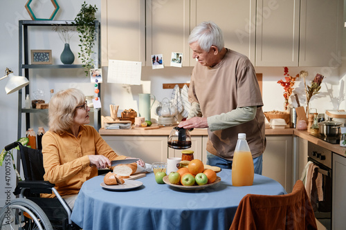 Senior woman cutting bread at the table with senior man pouring hot tea in cups during breakfast in the kitchen