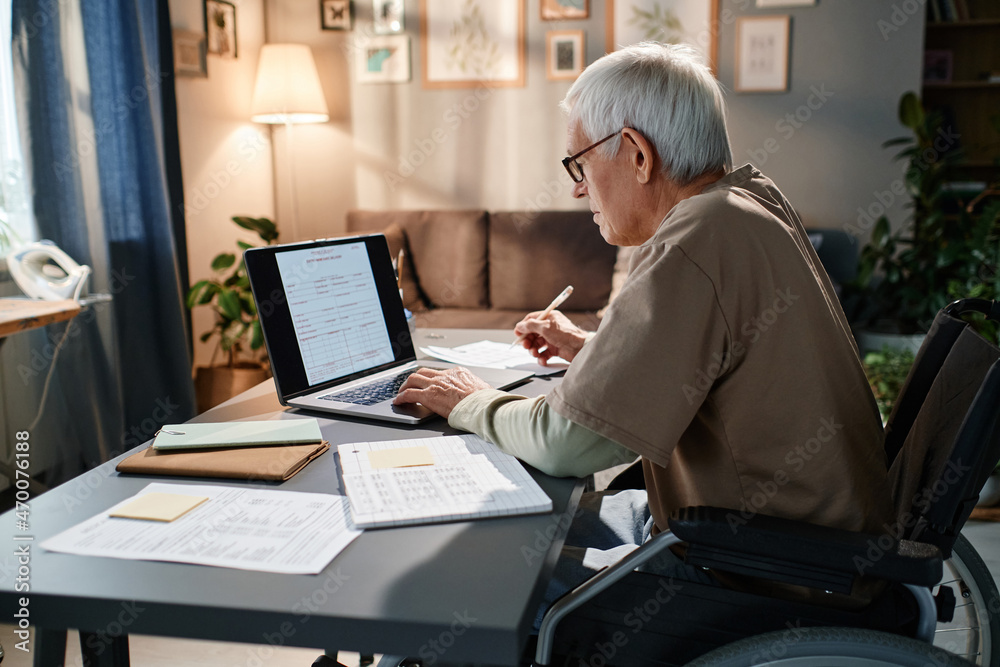 Senior man in eyeglasses using wheelchair sitting at the table and typing documents online on laptop, he working in the room Senior man in eyeglasses using wheelchair sitting at the table and typing documents online on laptop, he working in the room