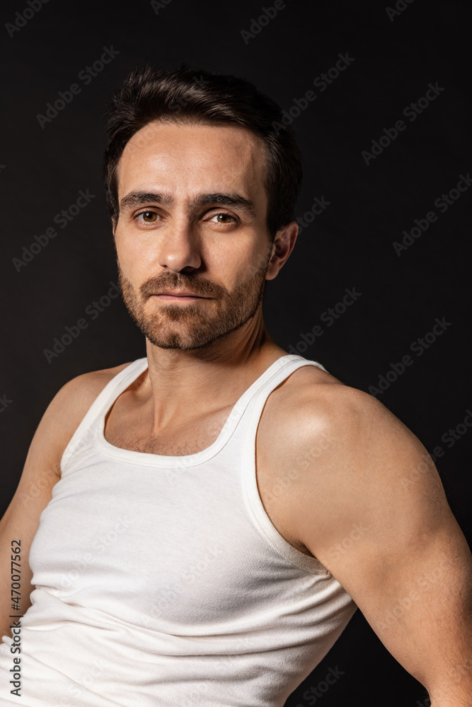 Close-up portrait of young handsome bearded man in white undershirt isolated on dark studio background. Emotions, facial expression Close-up portrait of young handsome bearded man in white undershirt isolated on dark studio background. Emotions, facial expression