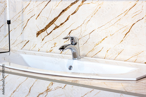 New modern metal water faucet with the white sink on the marble wall background in the bathroom.