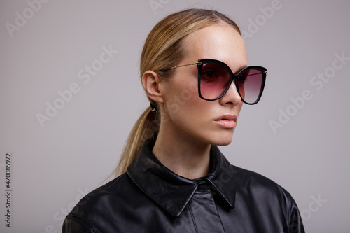 High fashion photo of a beautiful elegant young woman in a pretty black leather suit, bermuda shorts, jacket, stylish sunglasses posing on white background. Studio Shot. Slim figure.