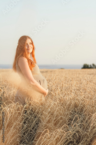 young pregnant woman walks in ripe wheat field at sunset, expectant mother with red hair relax in nature stroking her belly with her hand