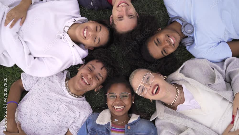 Relaxed teenager multiracial friends lying together on green grass with eyes closed, wake up and smile looking to camera Relaxed teenager multiracial friends lying together on green grass with eyes closed, wake up and smile looking to camera
