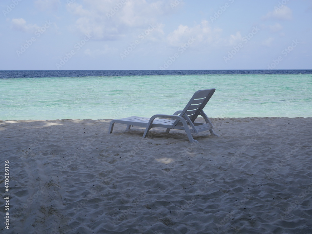 The seashore without people on a sunny summer vacation day. A white plastic sunbed stands in the shade of trees on the white sand of an ocean beach against the background of the sky and sea waves.