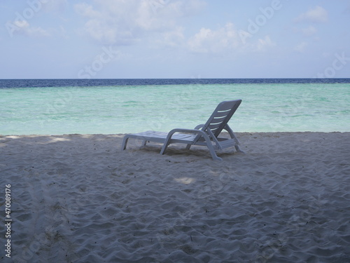 The seashore without people on a sunny summer vacation day. A white plastic sunbed stands in the shade of trees on the white sand of an ocean beach against the background of the sky and sea waves.