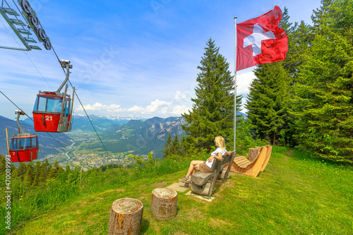 Red cable car cabin from Chur to Kanzeli and Brambruesch in Switzerland. Woman relaxing on top of Brambruesch. Swiss cable car of Chur or Coira with Swiss flag in Grisons canton.