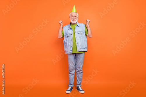 Photo of charming lucky retired man wear vintage jeans waistcoat birthday headwear rising fists smiling isolated orange color background