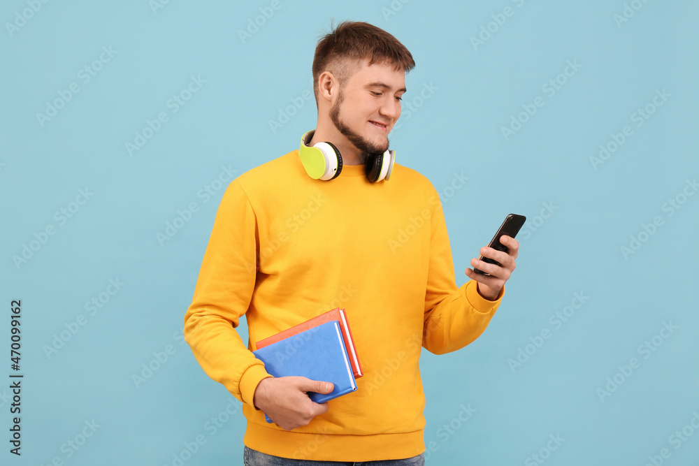 Young student with headphones and books using smartphone on light blue background