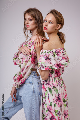 Two high fashion models in long dress with a red floral pattern, blouse, blue jeans. Beautiful young women. Studio shot, portrait. White background. Slim figure. Make up, hairstyle