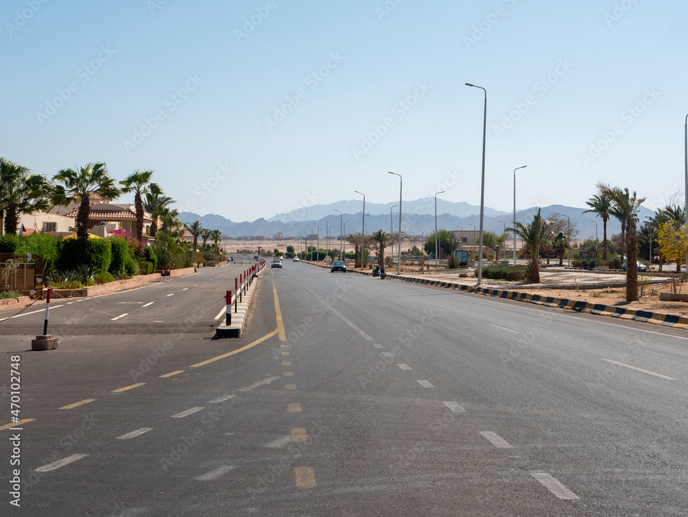 Fototapeta premium Safaga, Egypt - September 27, 2021: View of the main road stretching into the distance against the backdrop of mountains and blue sky. Cars drive along the highway to a neighboring city.