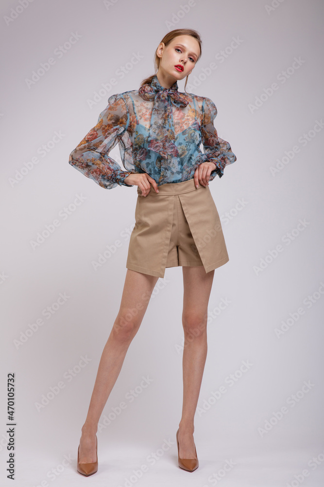 High fashion photo of a beautiful elegant young woman in a pretty blue transparent blouse with a floral pattern, a bow on the neck, short beige shorts similar to a skirt posing over white background High fashion photo of a beautiful elegant young woman in a pretty blue transparent blouse with a floral pattern, a bow on the neck, short beige shorts similar to a skirt posing over white background