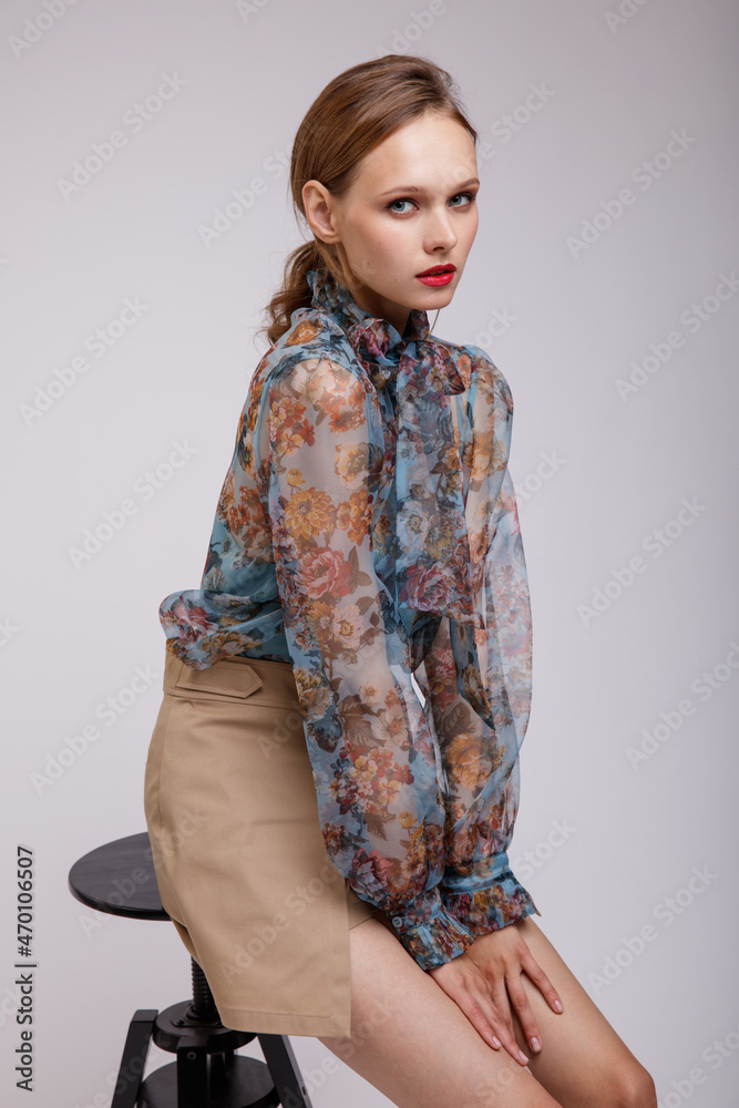High fashion photo of a beautiful elegant young woman in a pretty blue transparent blouse with a floral pattern, short beige shorts posing over white background. Model is sitting on a chair High fashion photo of a beautiful elegant young woman in a pretty blue transparent blouse with a floral pattern, short beige shorts posing over white background. Model is sitting on a chair