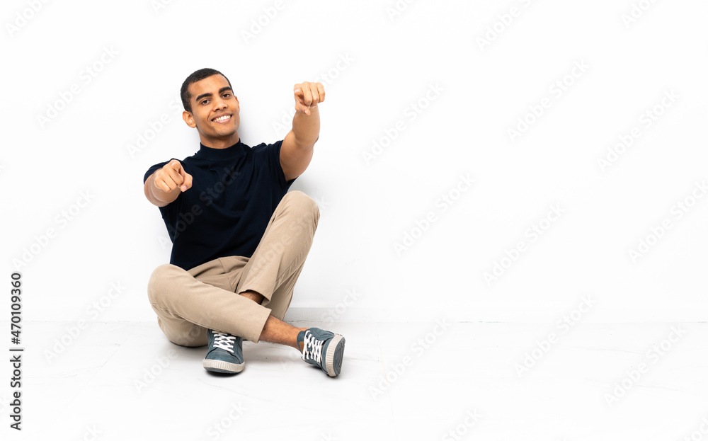Fototapeta premium African American man sitting on the floor points finger at you while smiling