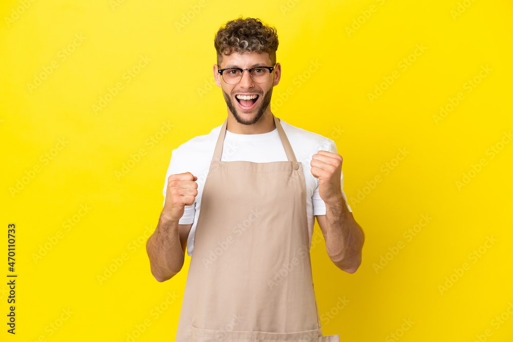 Restaurant waiter blonde man isolated on yellow background celebrating a victory in winner position Restaurant waiter blonde man isolated on yellow background celebrating a victory in winner position