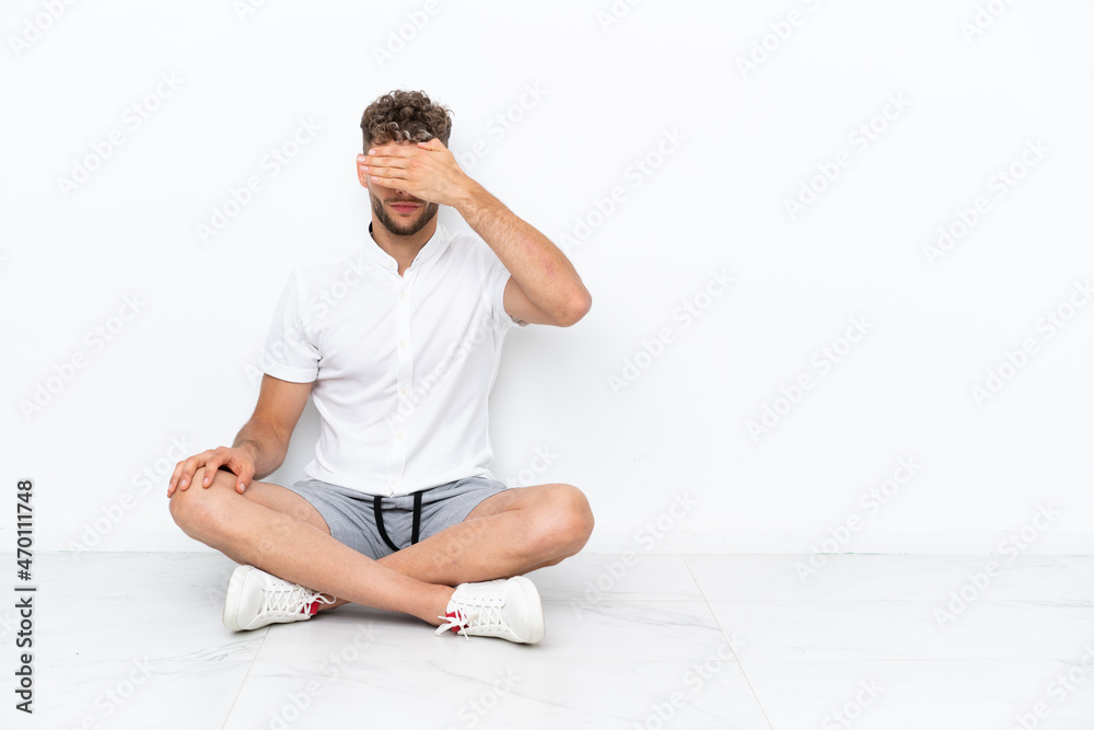 Young blonde man sitting on the floor isolated on white background covering eyes by hands. Do not want to see something Young blonde man sitting on the floor isolated on white background covering eyes by hands. Do not want to see something