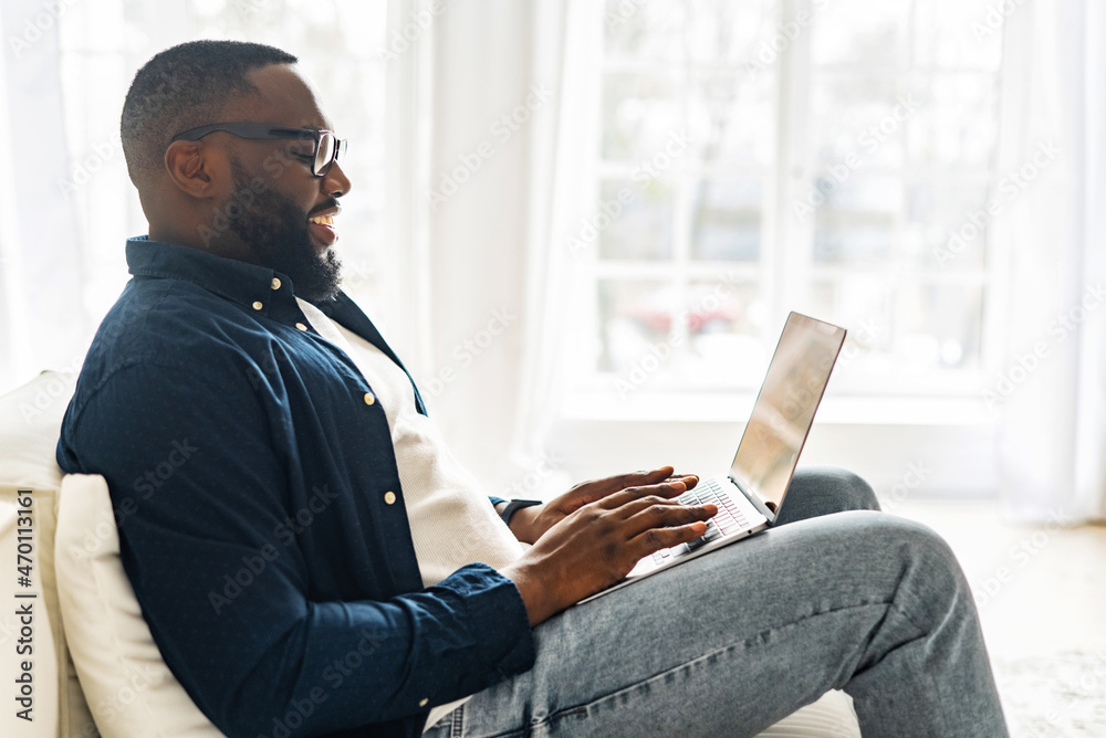 Side view at young modern African-American freelancer guy using laptop, black man in casual shirt and glasses typing sitting on the sofa, working remotely from home, web surfing, sending emails